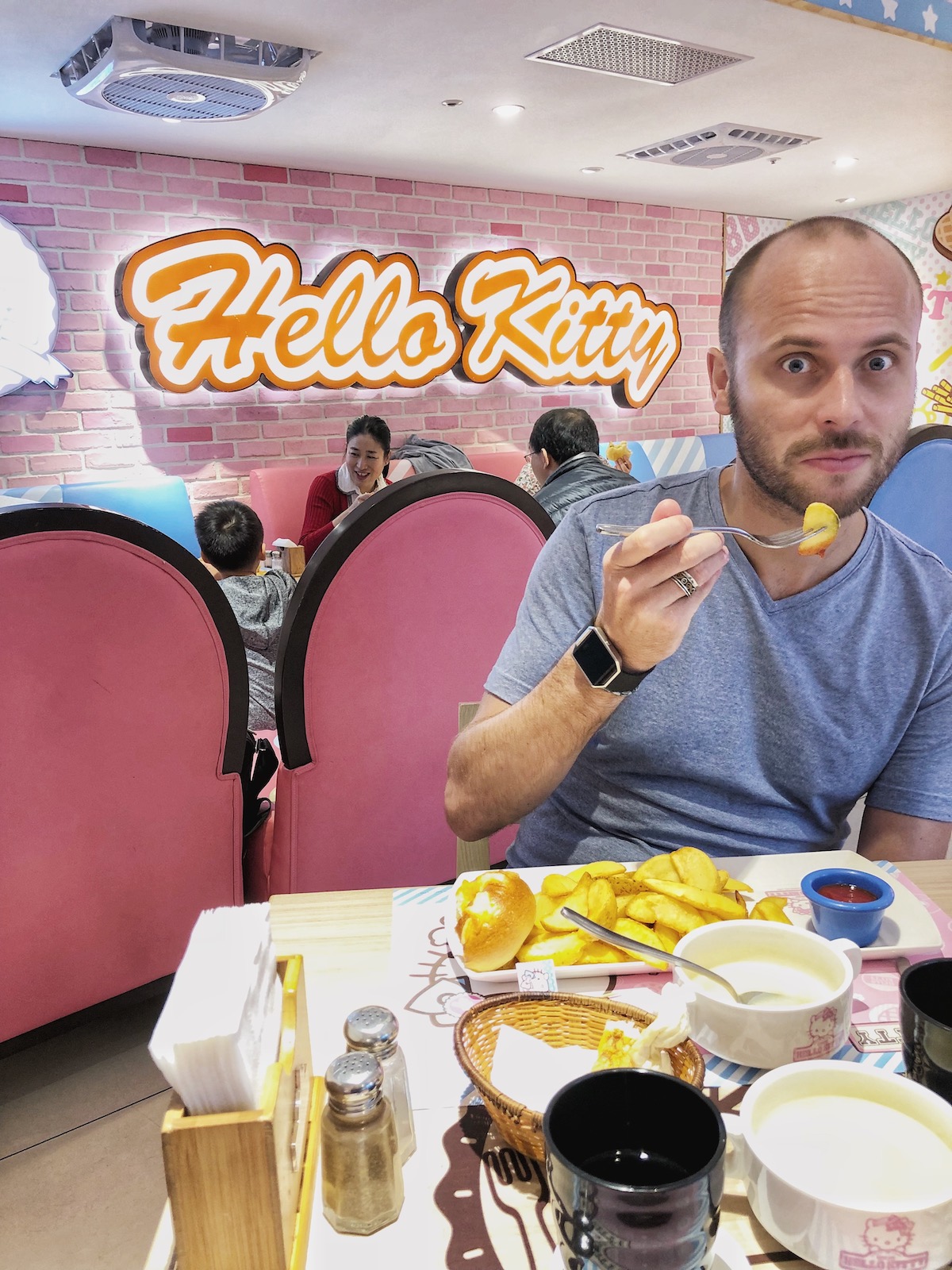 A man eats with a funny face in front of a bright Hello Kitty sign