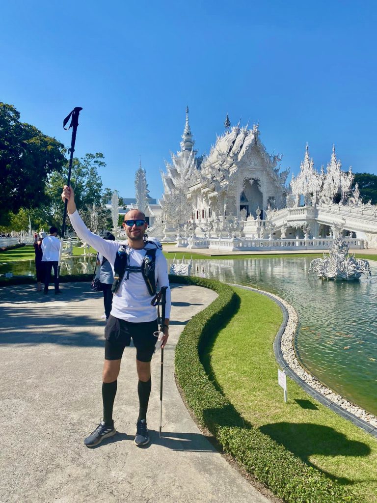 Man in sports gear raises arm aloft outside a white temple in Chiang Rai, Thailand.