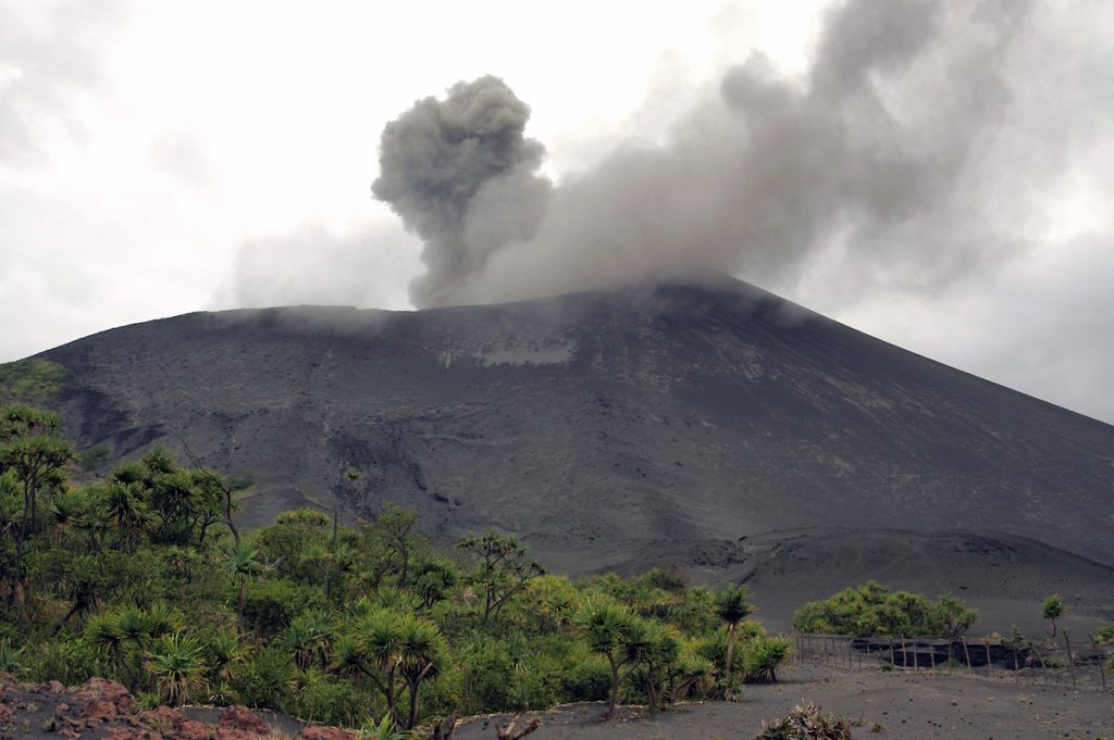 Green trees sit in front of smoke drifting from the top of a volcano.