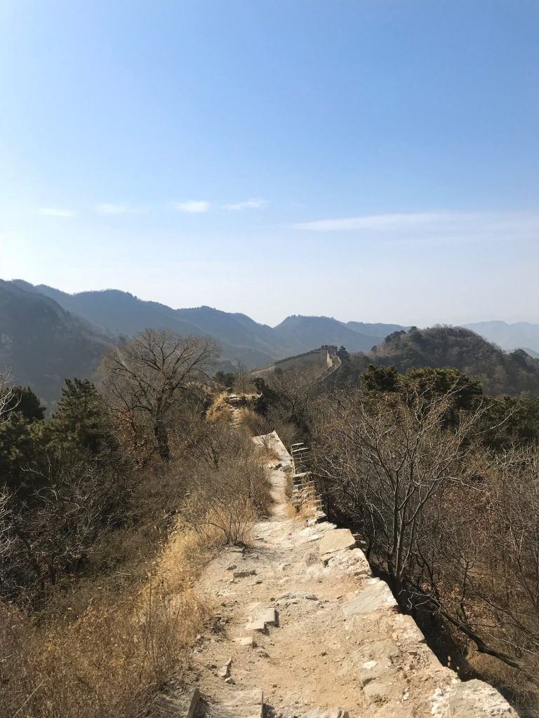 Broken pars of a long wall in China, with trees and mountains on either side.