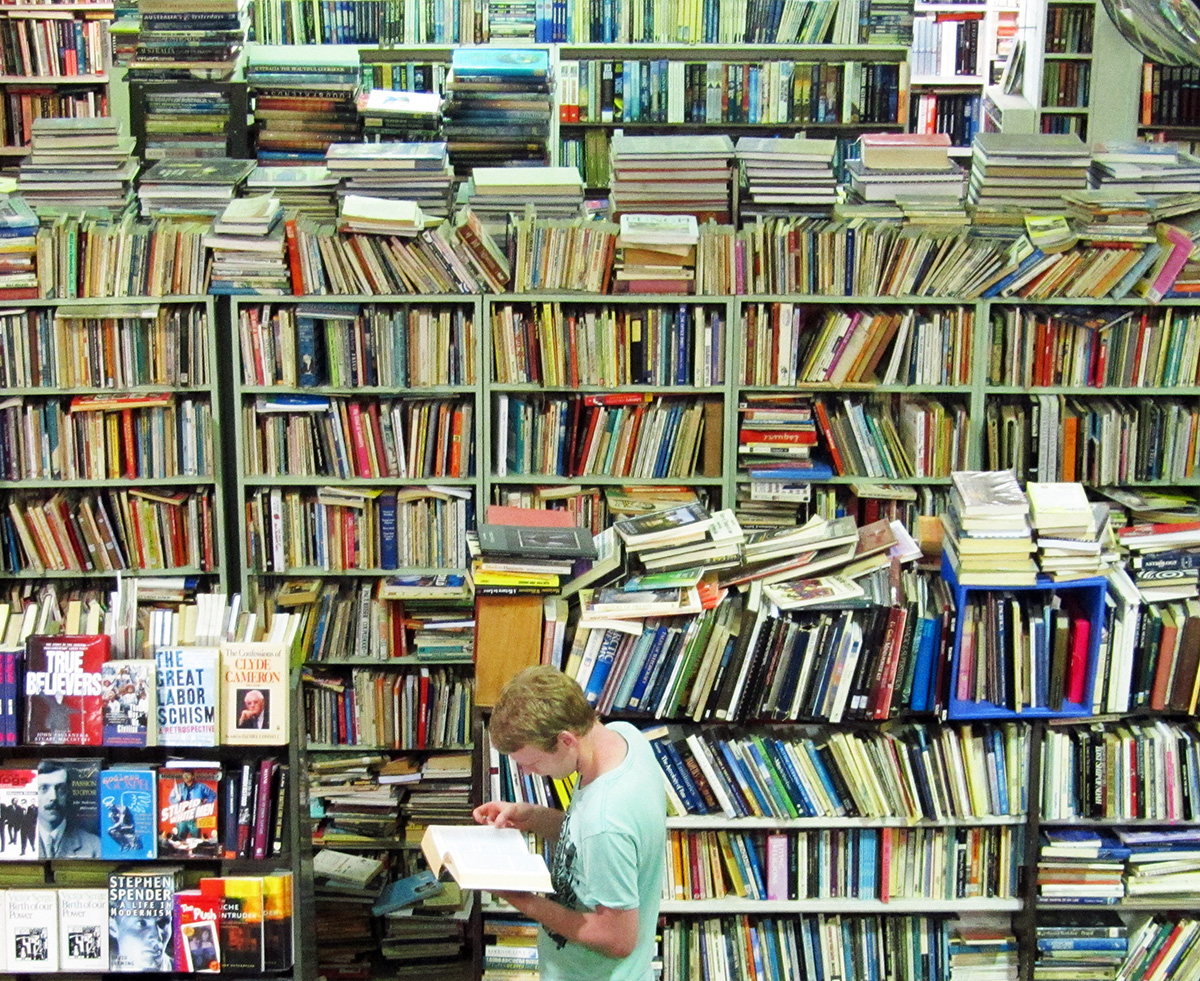 A young man reads an open book in a huge room full of mountains of books stacked high.