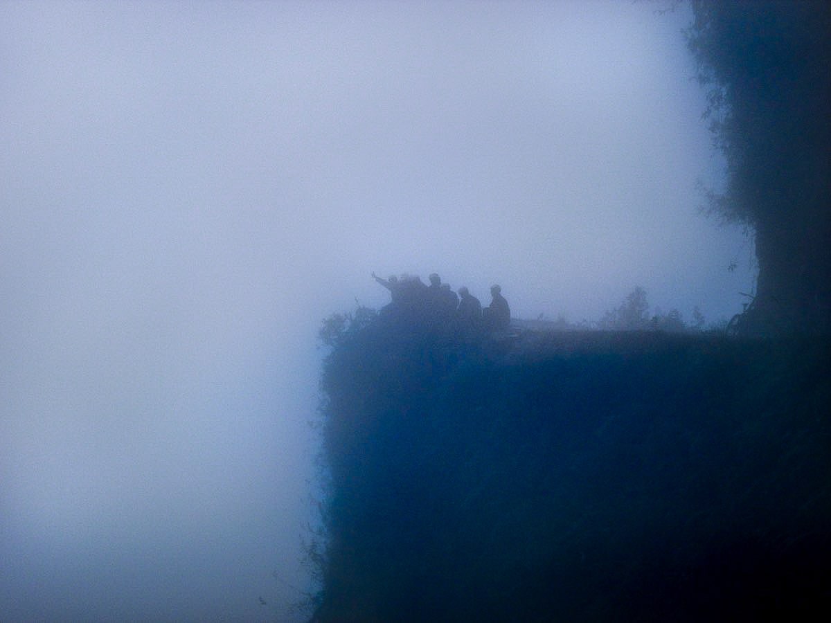 People wave in the mist of Bolivia's Death Road while sitting on a cliff.