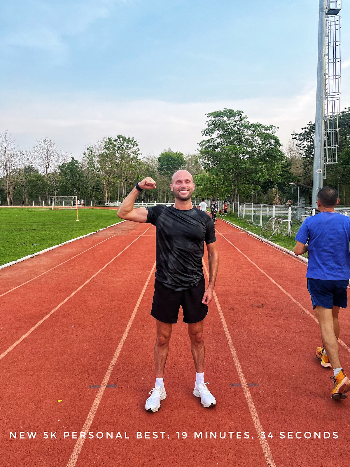 A smiling man in black t shirt and shorts tenses his biceps and smiles on a running track