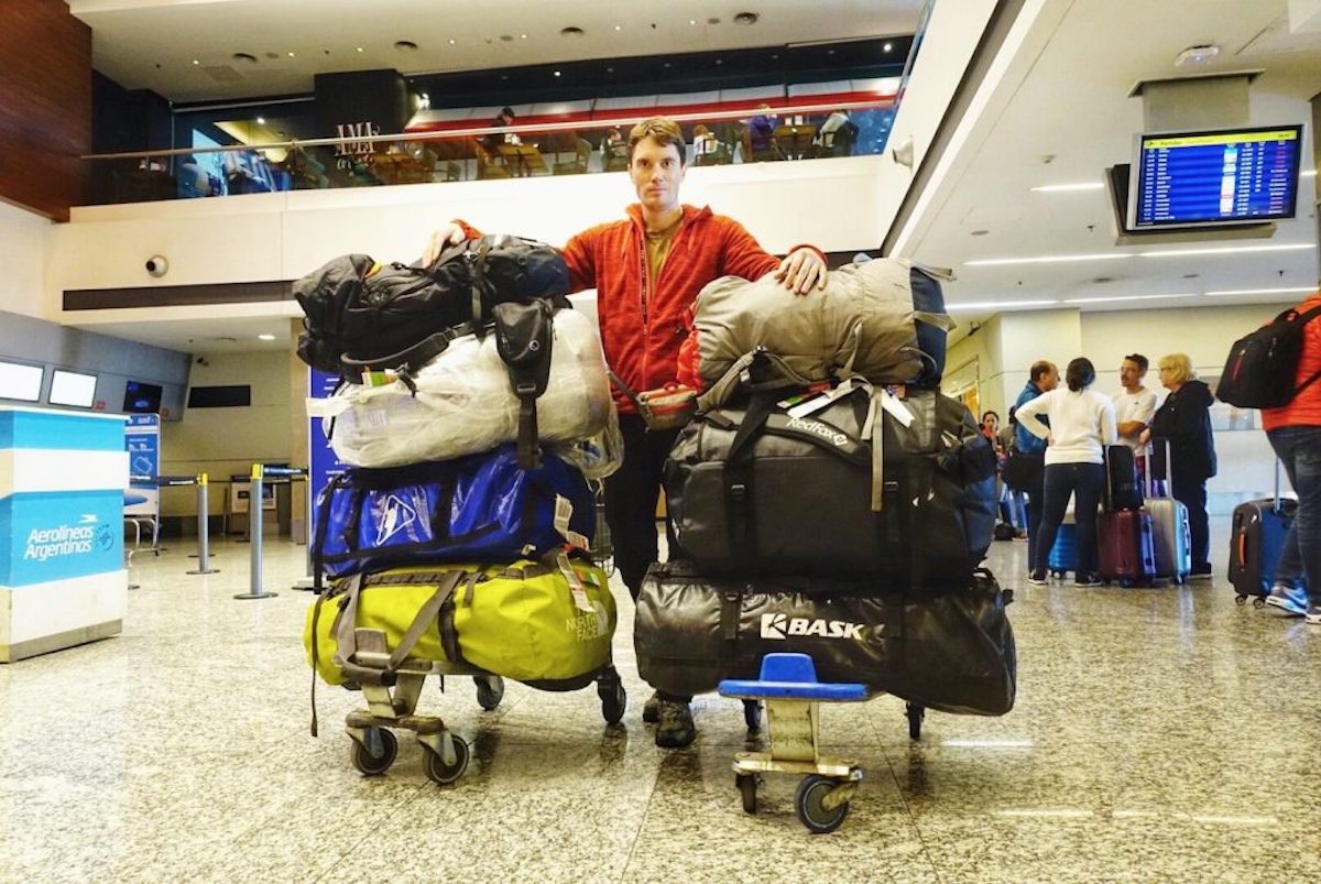 A man in an orange jacket stands in the airport between two large luggage trolleysn packed with mountain bags stacked on top of one another