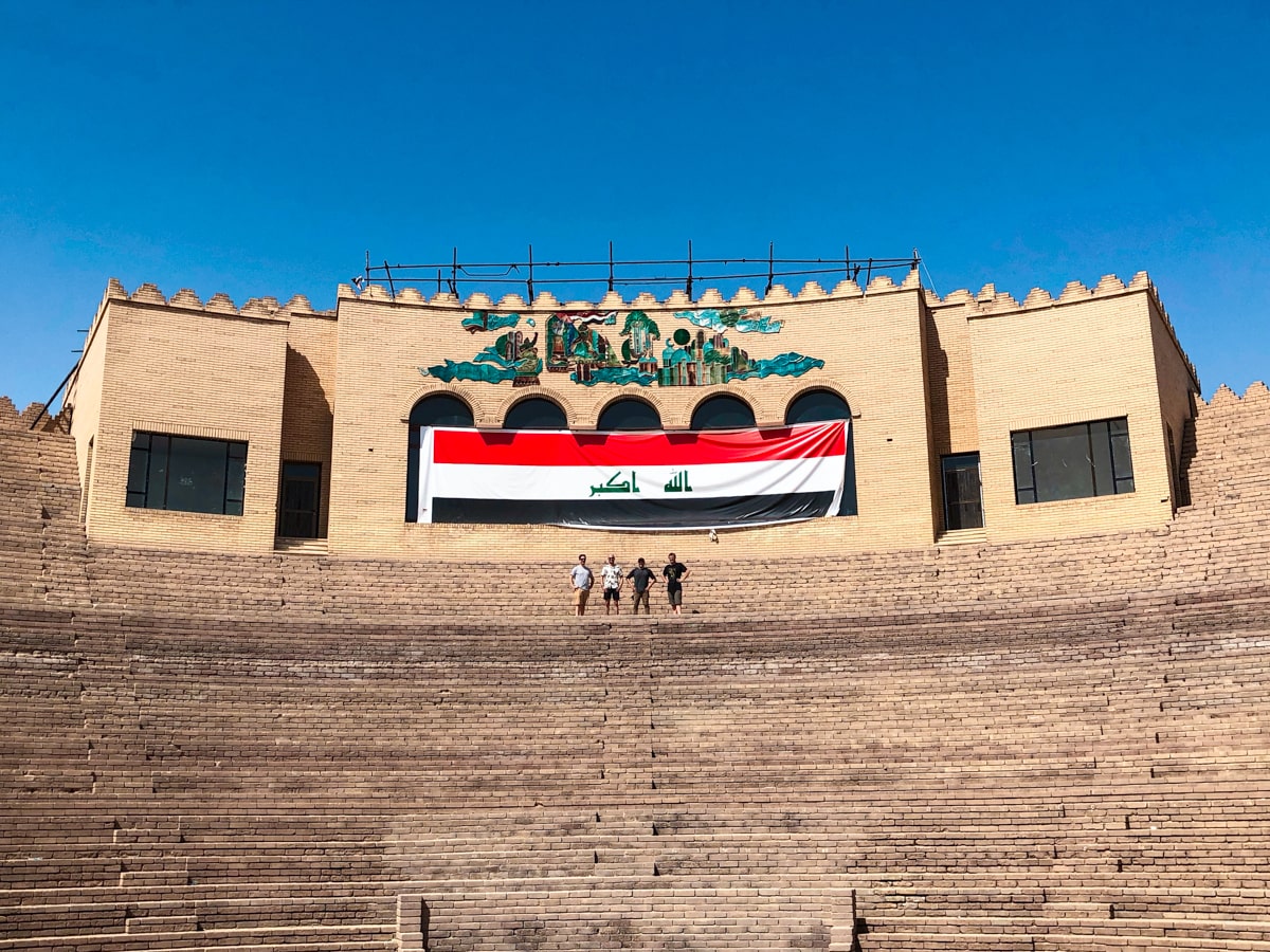 Men stand in the Amphitheatre of Ancient Babylon in Iraq, the Iraq flag is draped over the top of the stairs