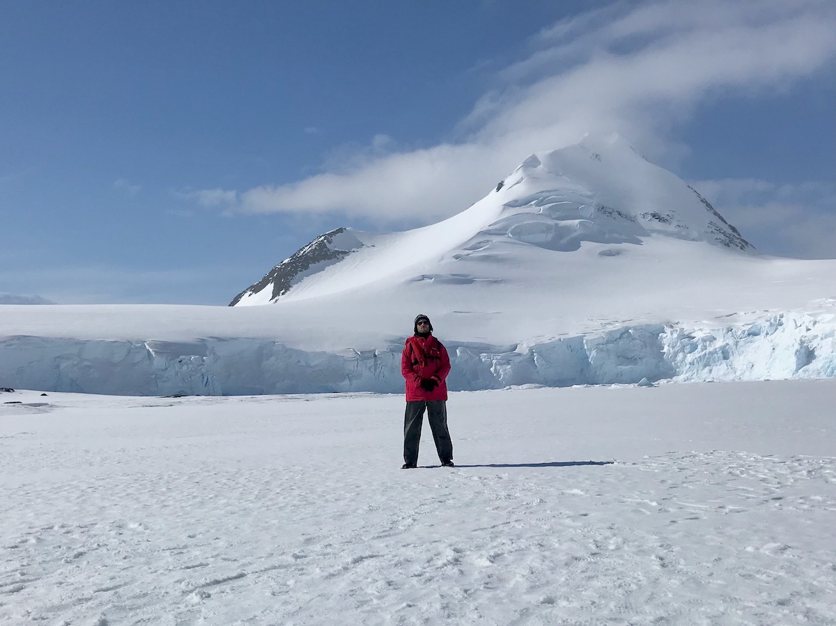 Man in red parka and grey hiking pants stands in the snowy landscape of Antarctica