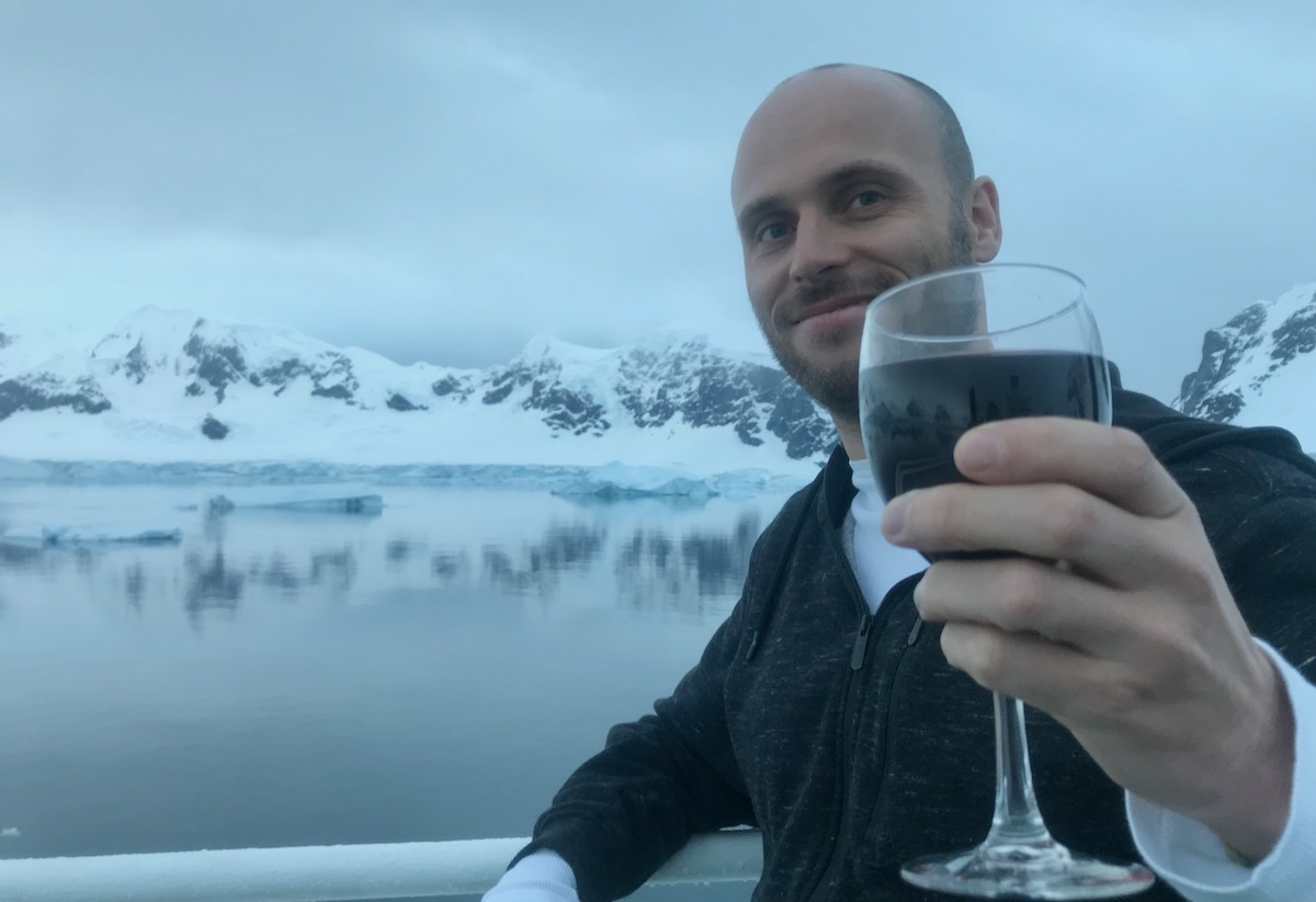 A man raises a glass of red wine near a glacier which reflects off a body of water