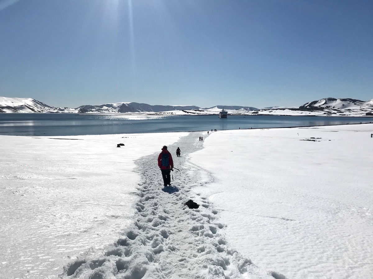 People individually walk on a trail in the snow towards a blue lake during a trip to Antarctica
