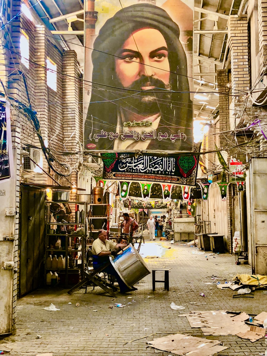 A workman crafts a large metal tin in Baghdad Souk with a large portrait of a bearded Arabic man strikingly hangs above him