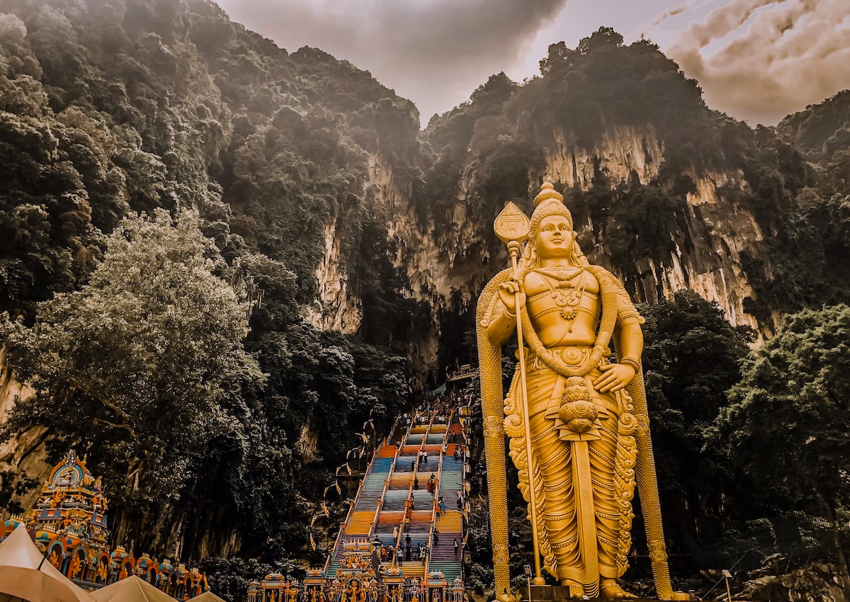 A large, majestic golden Lord Murugan stands tall in front of multicoloured steps and large limestone caves in Kuala Lumpur, Malaysia