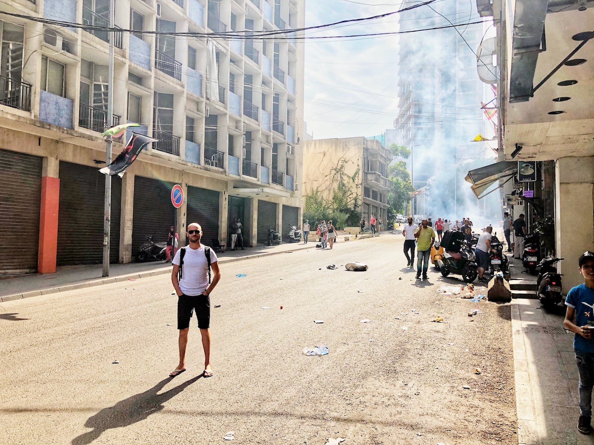 A tourist in a whhite t shirt and black shorts and sunglasses stands in the middle of the street during a mass protest in Beirut, Lebanon