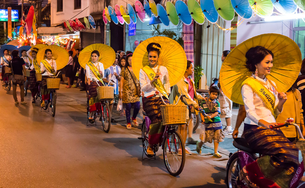 Pretty Thai ladies drive bicycles while holding large umbrellas in one of the best festivals in Thailand.