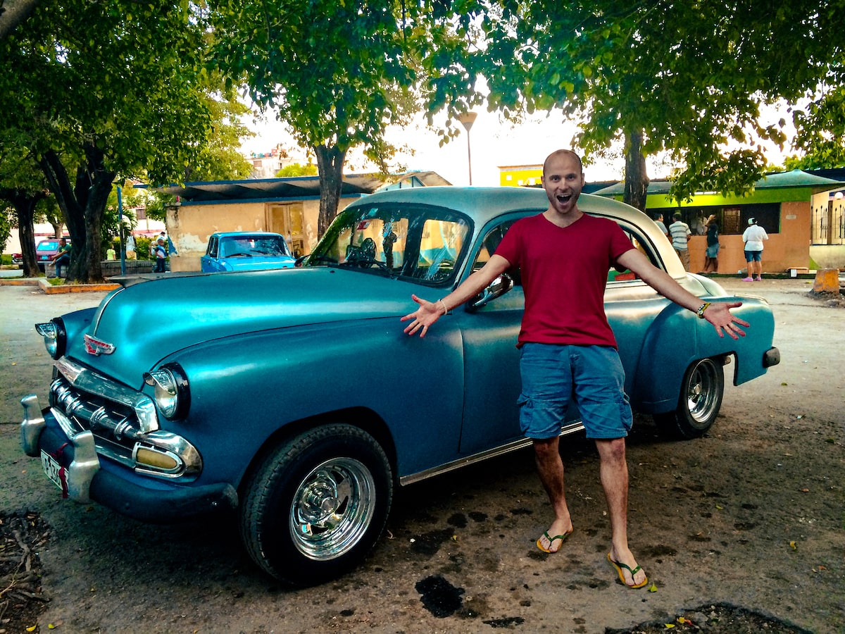 A male tourist in a red t-shirt stands outside a blue classical car in Havana, Cuba.