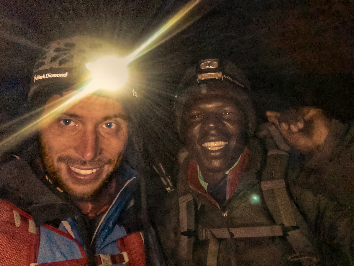 Two men smile at the camera up close with head torches on their foreheads. 