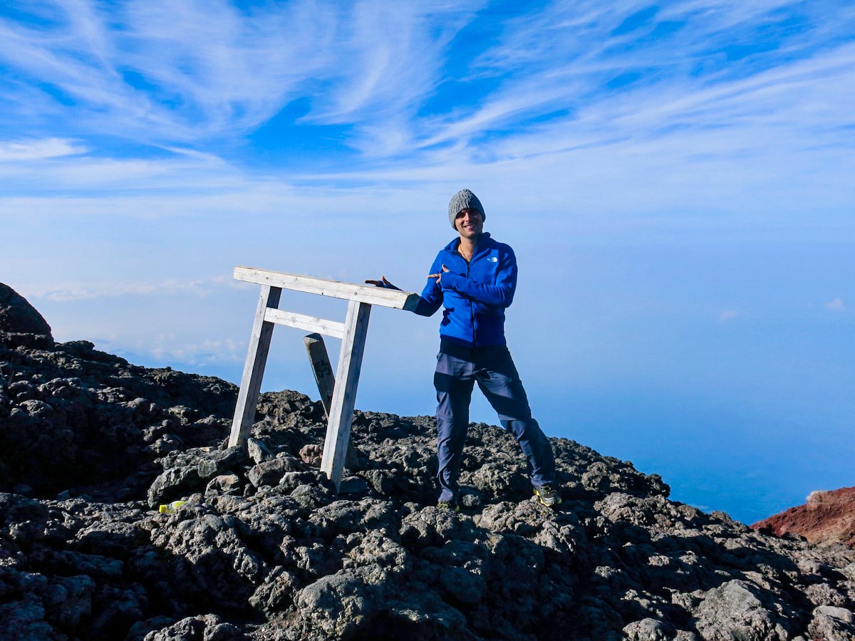 A man in blue smiles and points as he summits Mount Fuji in Japan.