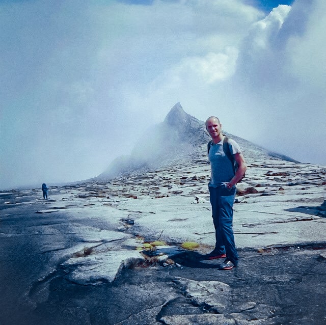 Man poses next to summit of a mountain with the backdrop of clouds