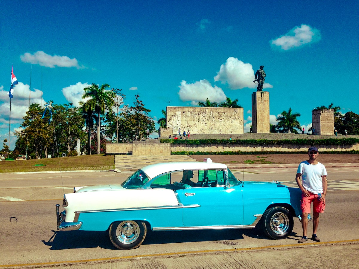 A male tourist poses beside a blue and white Chevrolet car outside the Che Guevara monument in Santa Clara, Cuba.