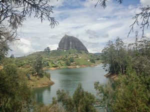 El Peñol Rock, a large rock outside of Medellin sits in a large body of water amongst greenery during the day
