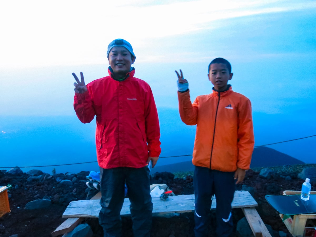 A Japanese father and son pose on a mountain with the one-handed piece sign. 