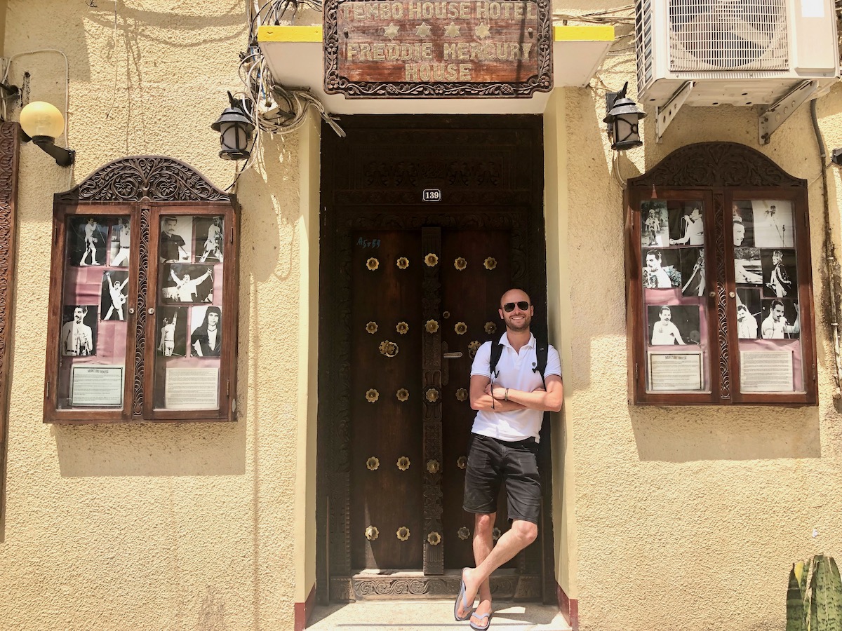 A man in sunglasses smiles as he leans against a yellow building with pictures of Freddie Mercury