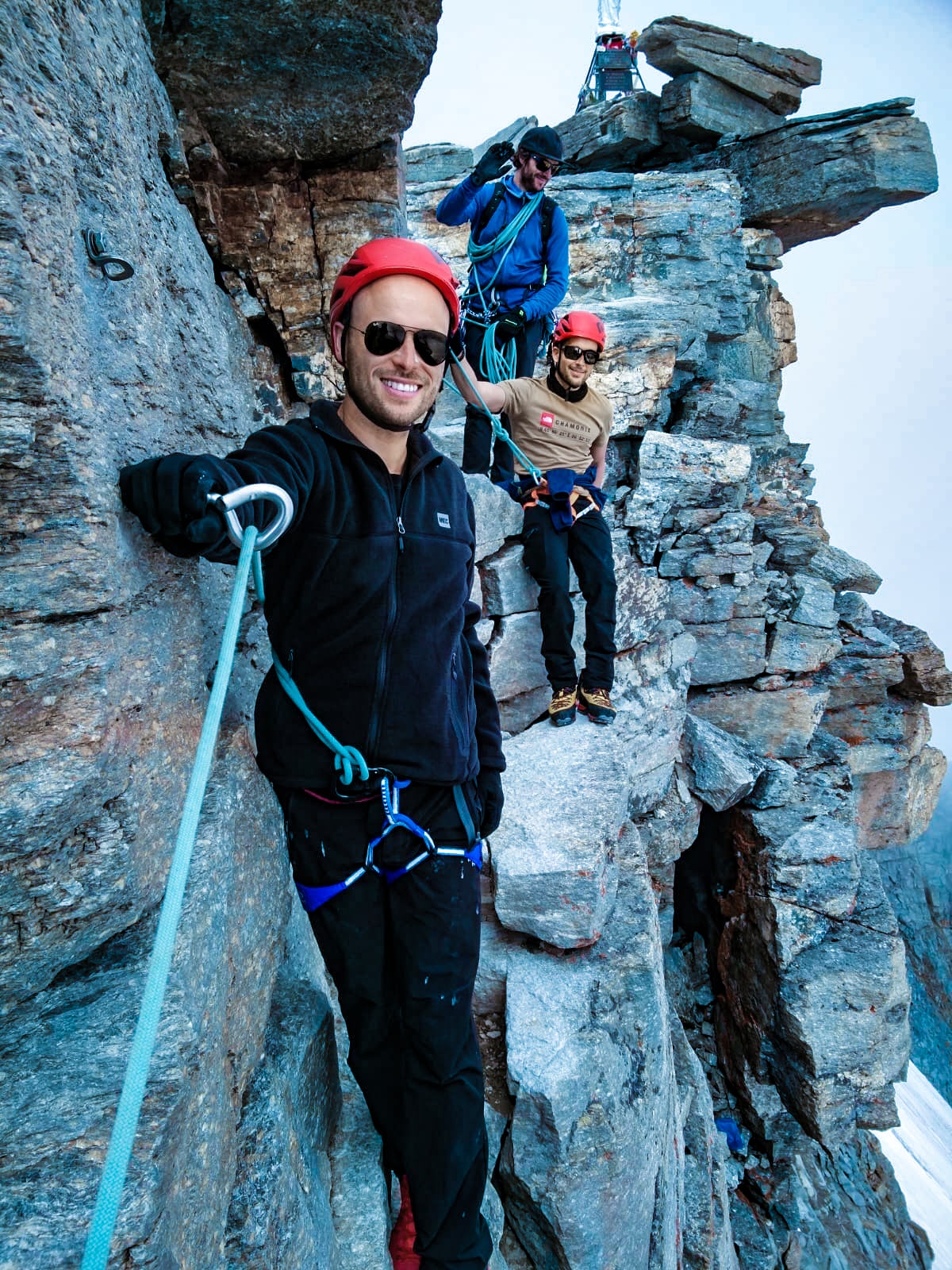 Three men pose for a smile on a mountain with jagged rocks, all of them roped in to one another.