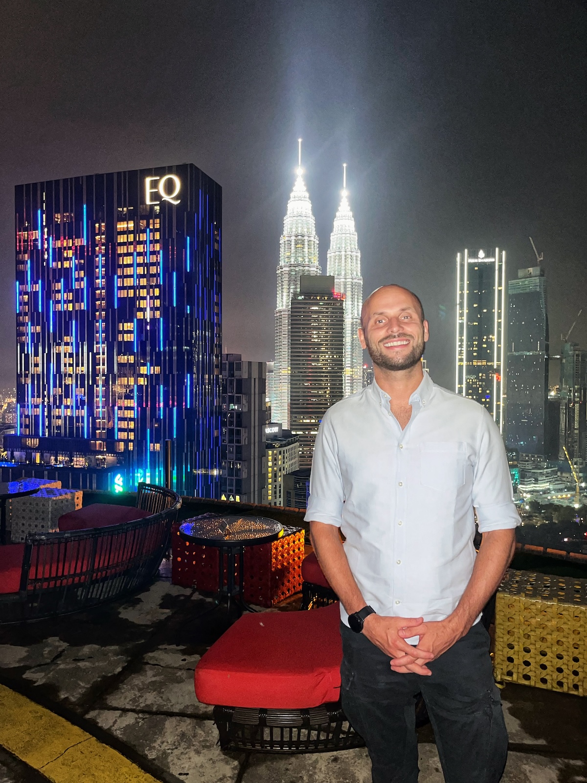 A man standing on the rooftop of Helibar in Kuala Lumpur at night, with the illuminated Petronas Twin Towers and other skyscrapers in the background. The vibrant city lights create a stunning urban skyline.