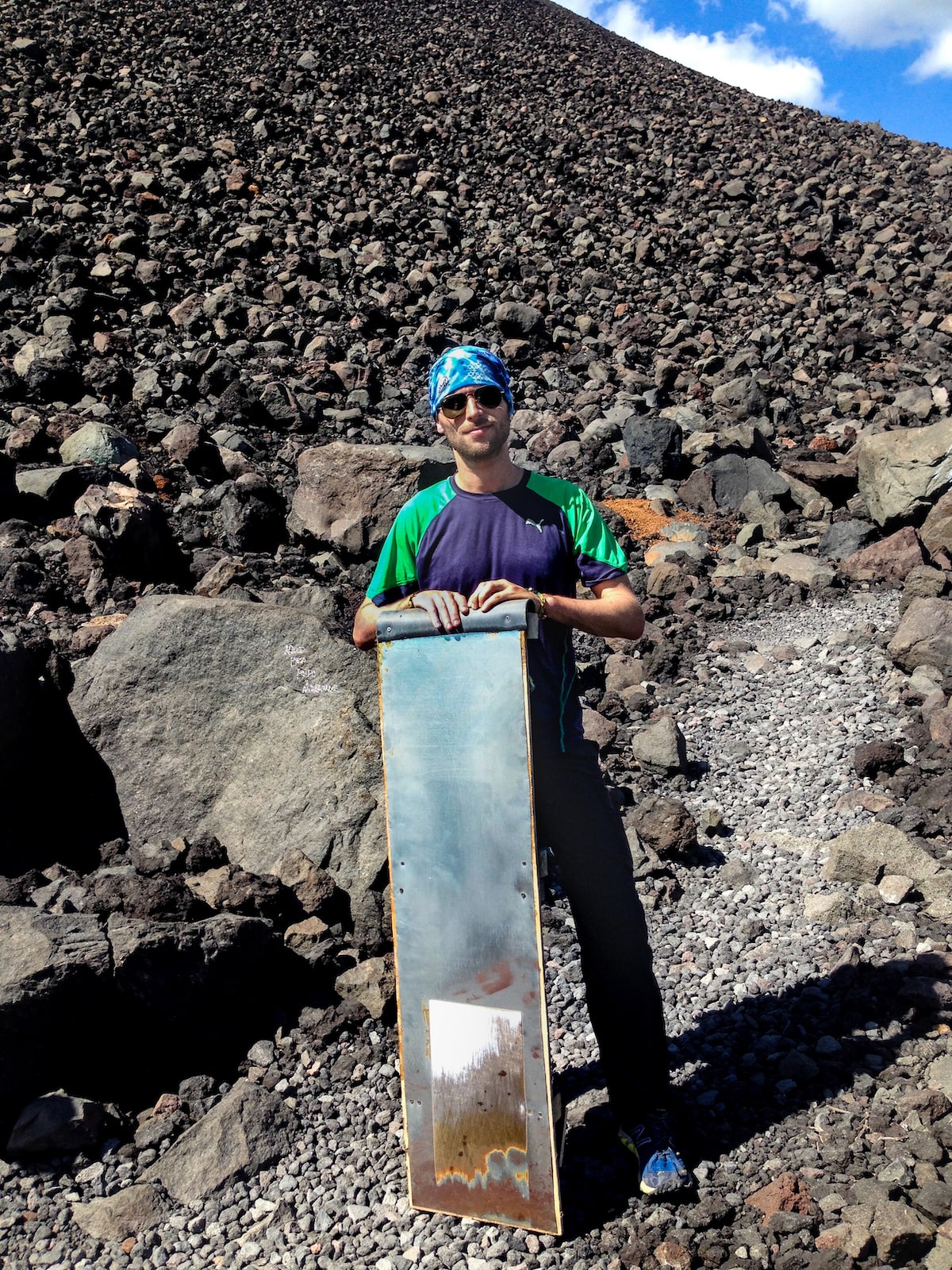 A male tourist in sunglasses and a blue bandana poses with a volcano board at the bottom of Cerro Negro, Nicaragua.