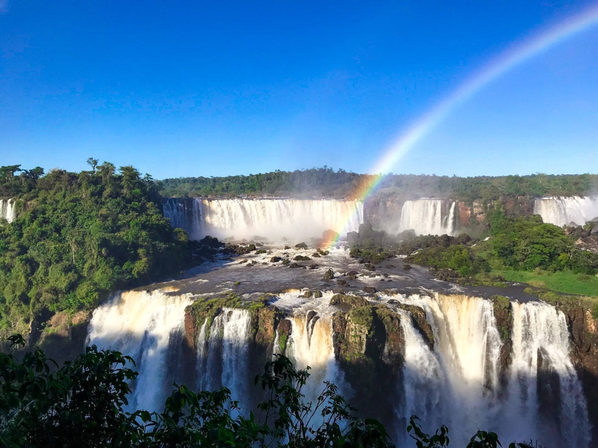 A rainbow shines over a slew of waterfalls on a sunny day from The Iguazu Falls Brazil side  