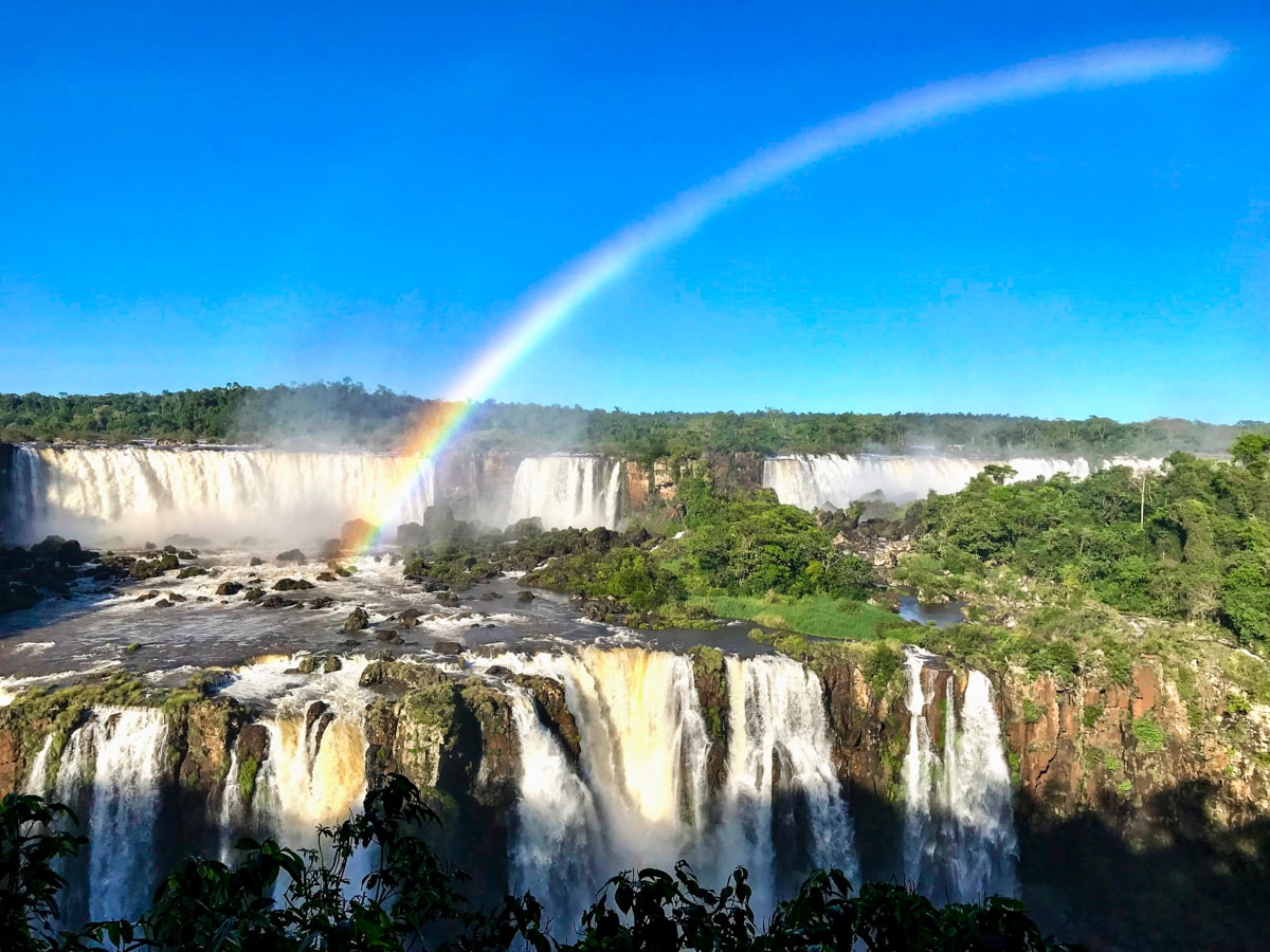 A rainbow shines over a gigantic waterfall, giving a treat to tourists visiting Iguazu Falls