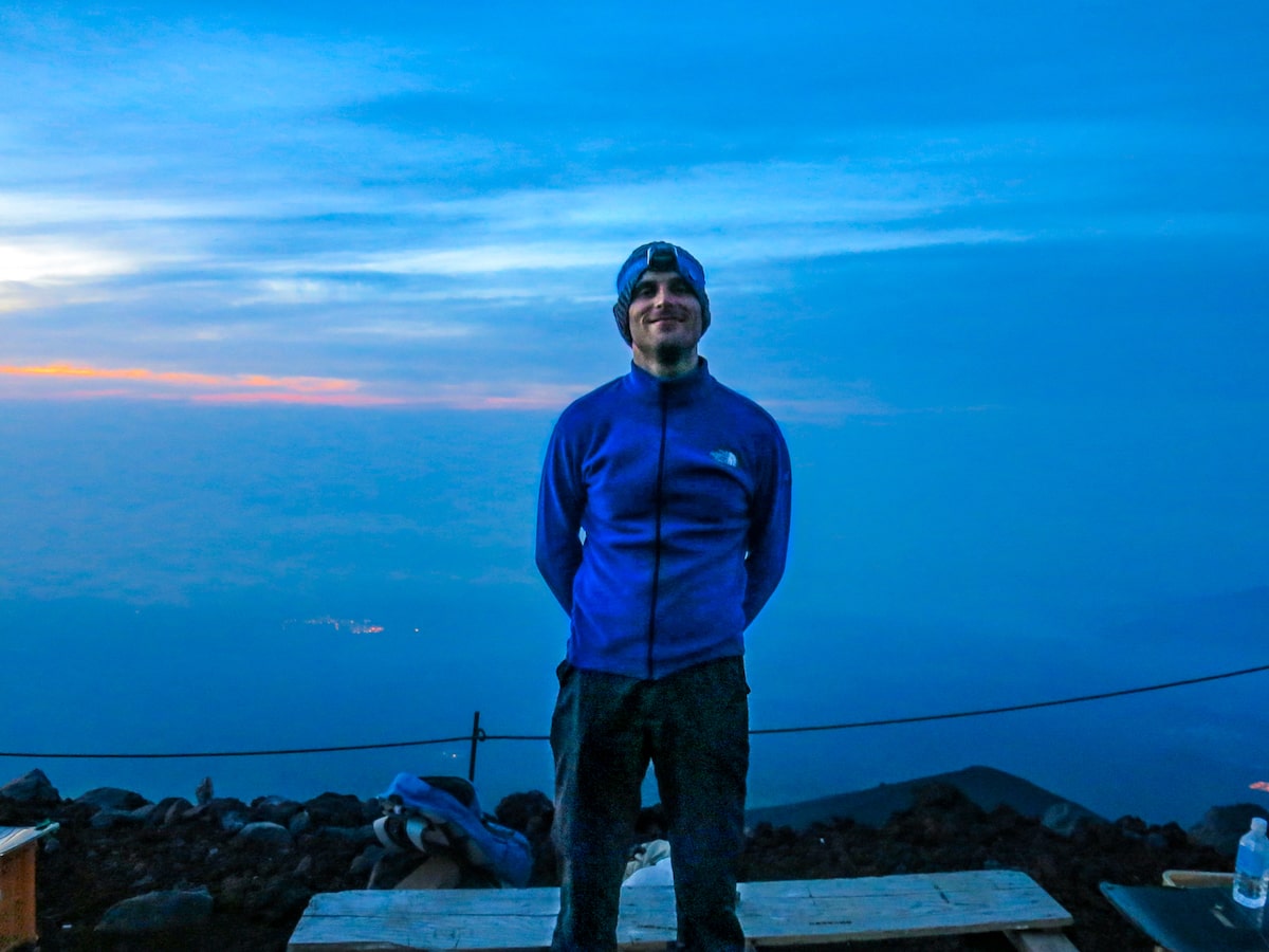 A man in blue with a headtorch smiles on a mountain at dusk.