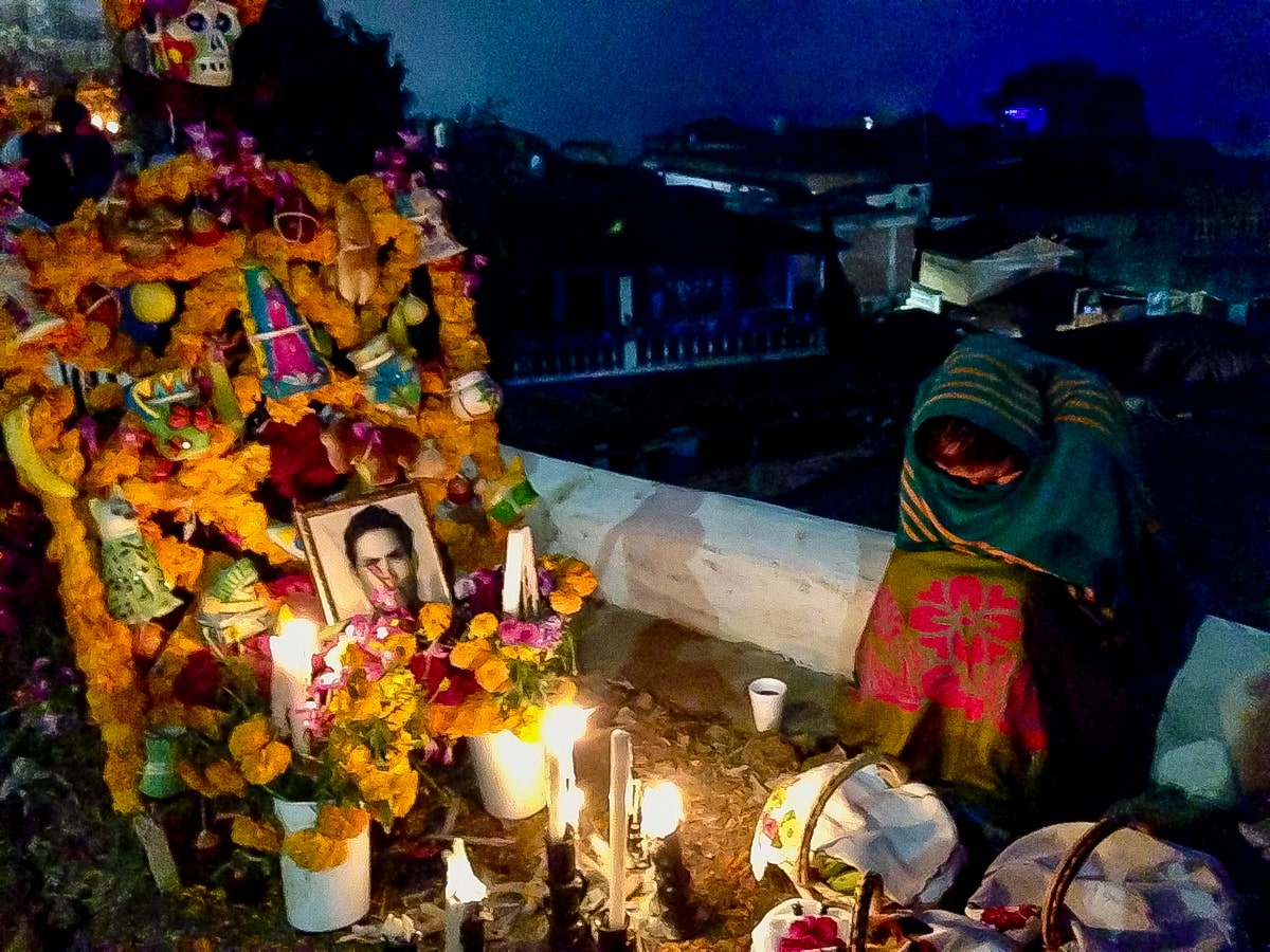 A lady wrapped in a scarf lowers her head next to a decorated grave, with a photo of a young man.