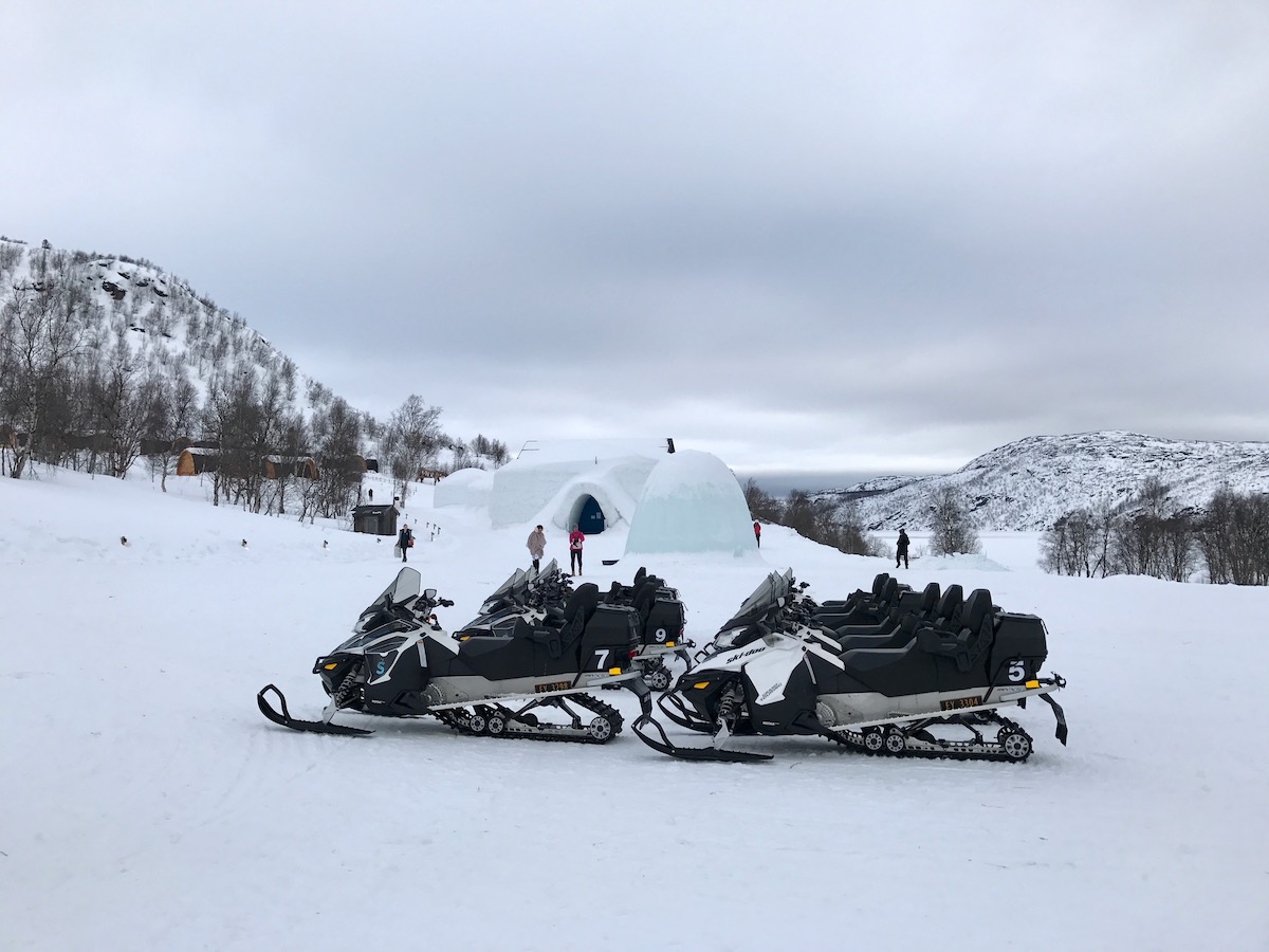 An igloo and snow hotel surrounded by snowy mountains with snowmobiles parked in front