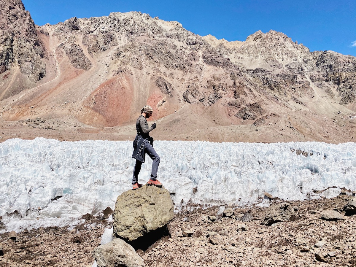 A mountain climber checks his sunglasses on Aconcagua while standing on a rock near glacier ice, with brown mountain landscapes and a bright blue sky behind him