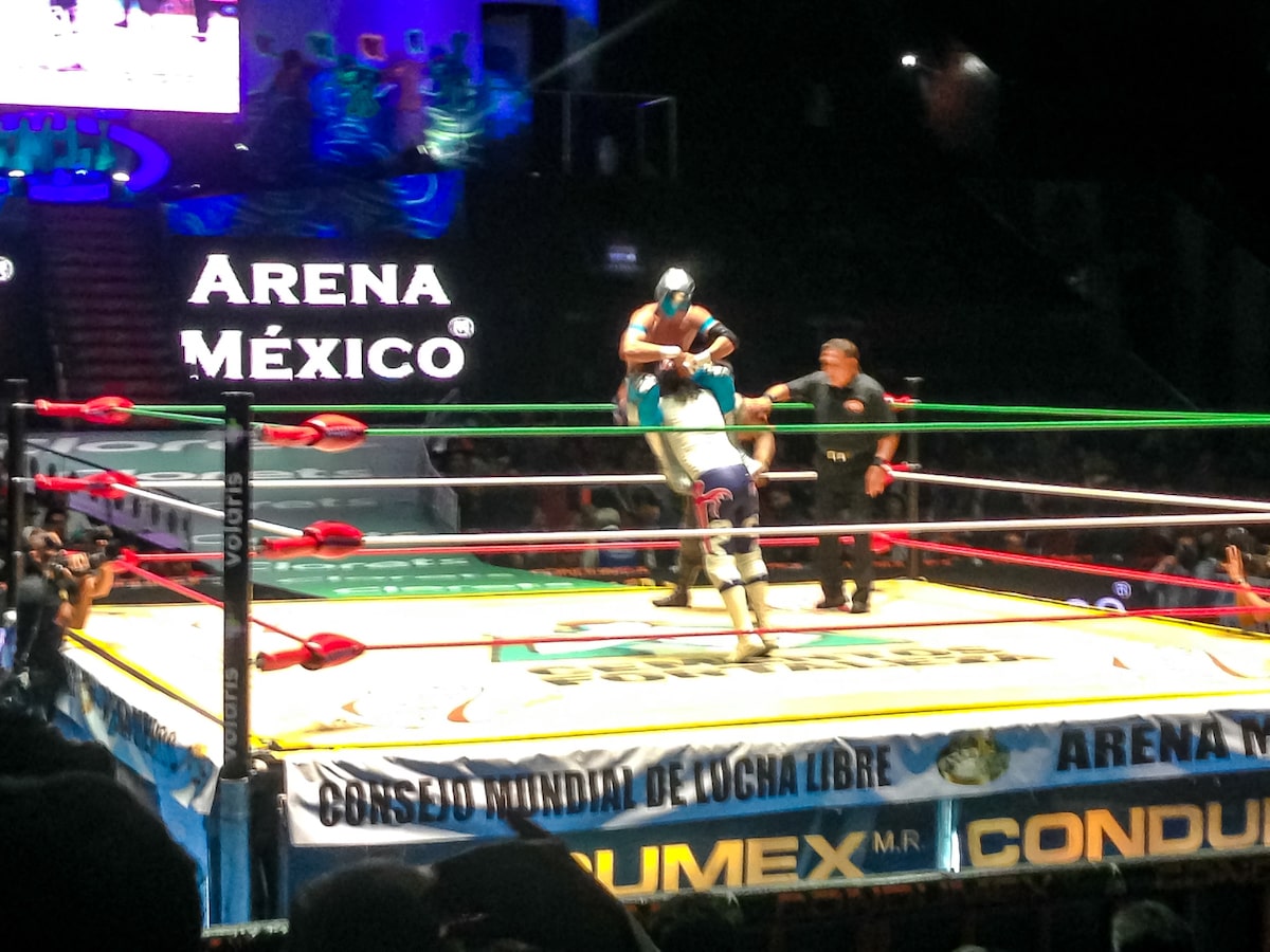 A wrestler picks up a masked wrestler in a wrestling ring in a packed arena, in Mexico City.