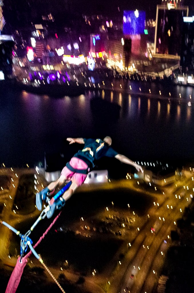 A man in a black t shirt and pink shorts bungee jumps off a large tower in Macau, China.