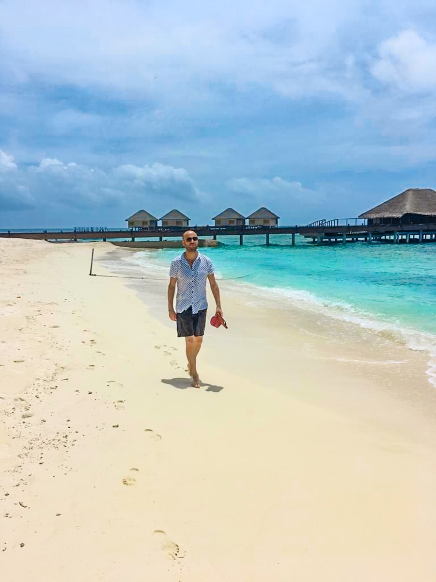 A man in a white shirt and blue shorts walks along the beach in a Maldives resort