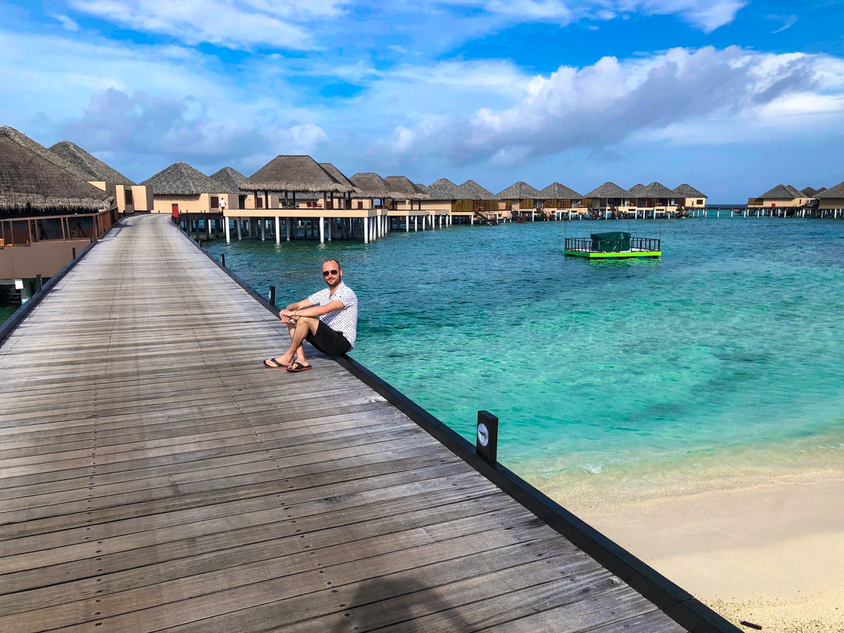 A tourist in the Maldives sits on a wooden walkway with the backdrop of water villas and beautiful blue sea