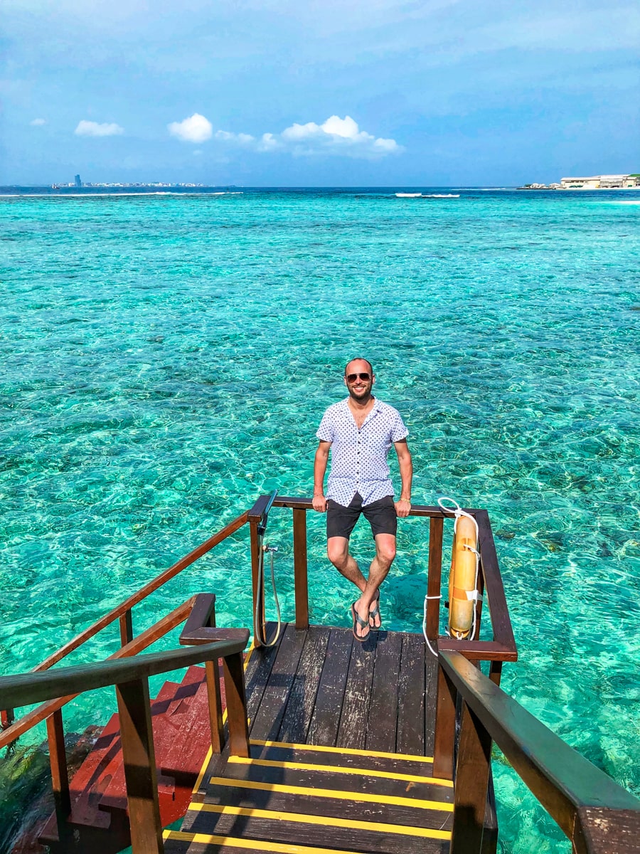 A male tourist with sunglasses smiles on a balcony with the clear blue sea in Maldives 