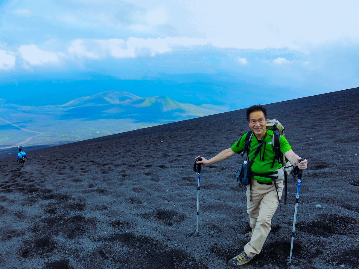 A Japanese man smiles with walking poles on Mount Fuji.