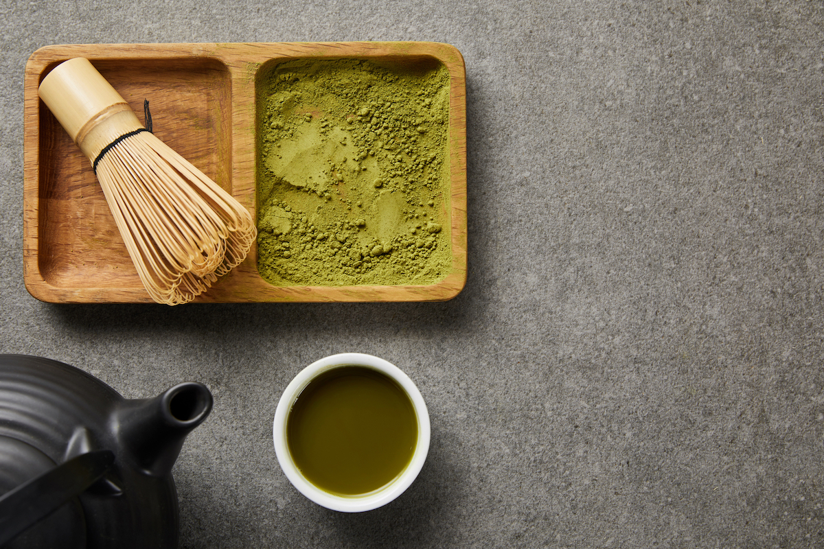 Top view of bamboo whisk and matcha powder on board near a black teapot