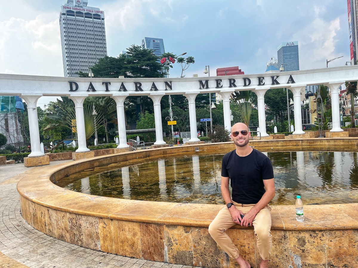 A man sitting on the edge of a circular fountain in front of the Dataran Merdeka sign in Kuala Lumpur, Malaysia, with tall buildings and lush greenery in the background