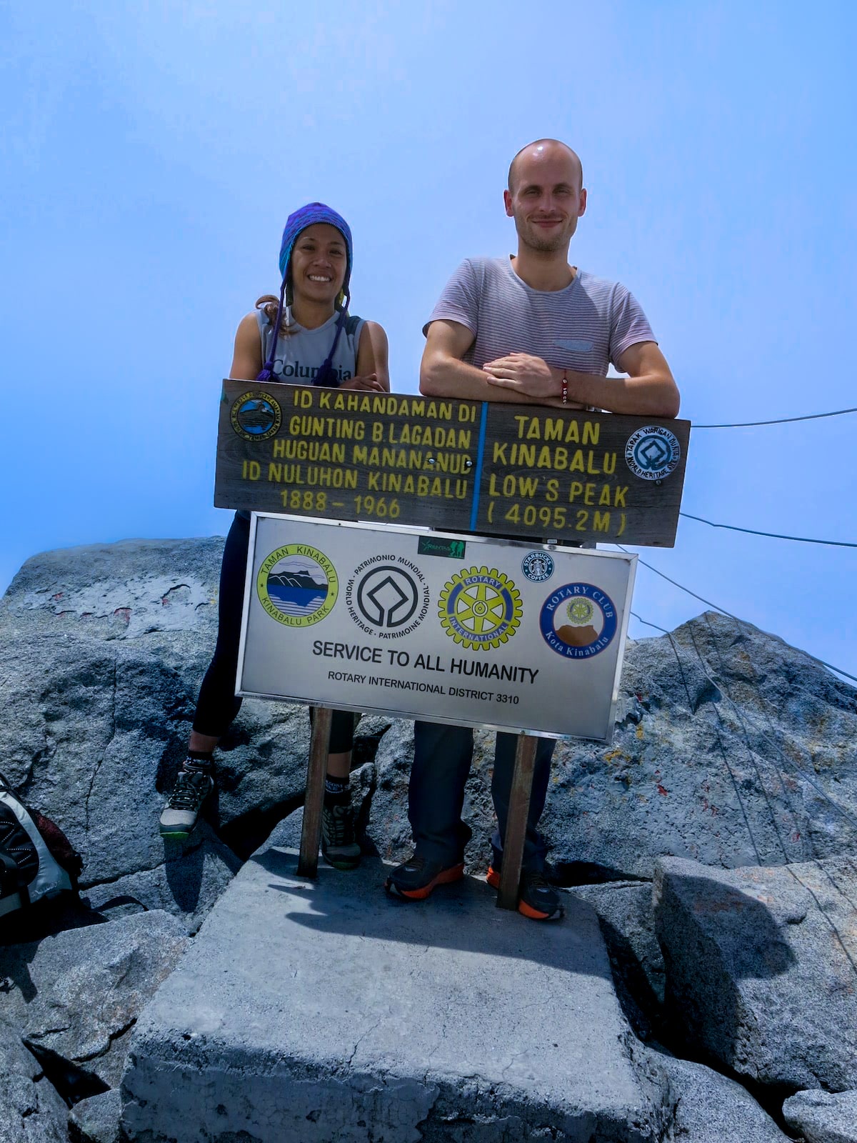 A man and a woman smile as they lean on a signpost on top of a mountain.