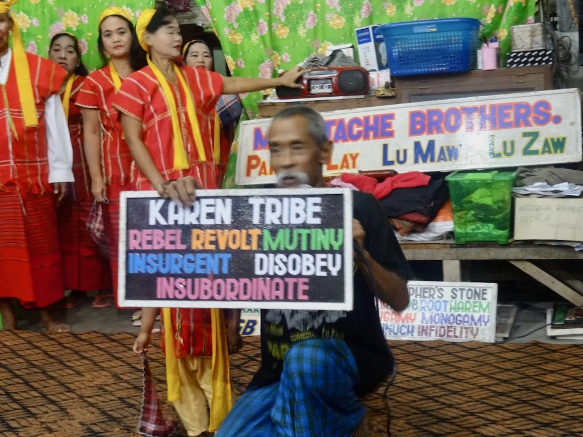 A Burmese man holds up a sign with multiple words, 5 women stand behind him dressed in orange 