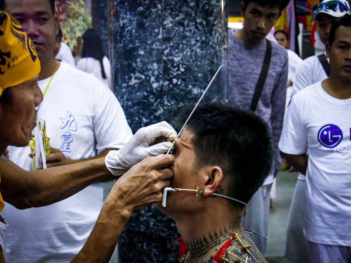 A man with a mini dagger through his ear has his eyebrow pierced with a large needle. 