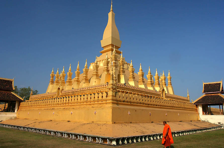 A monk in orange walks past a beautiful large golden temple in Vientiane, Laos