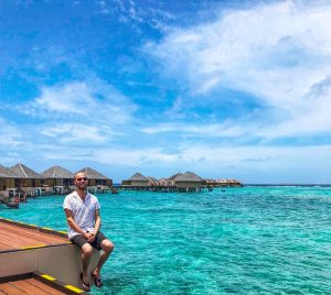 A male tourist in black shorts and a white short poses in front of water villas in The Maldives