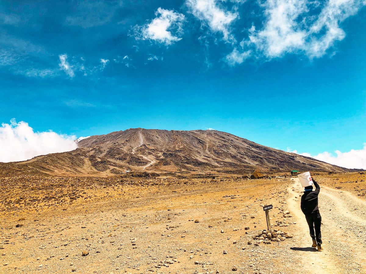 A man walks towards a mountain with a bucket on his head.