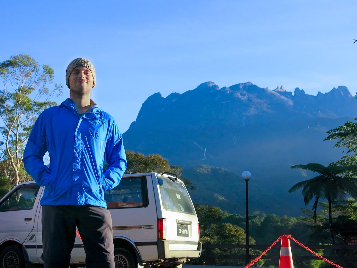 A man in a grey hat and blue jacket poses at the bottom of a mountain. 