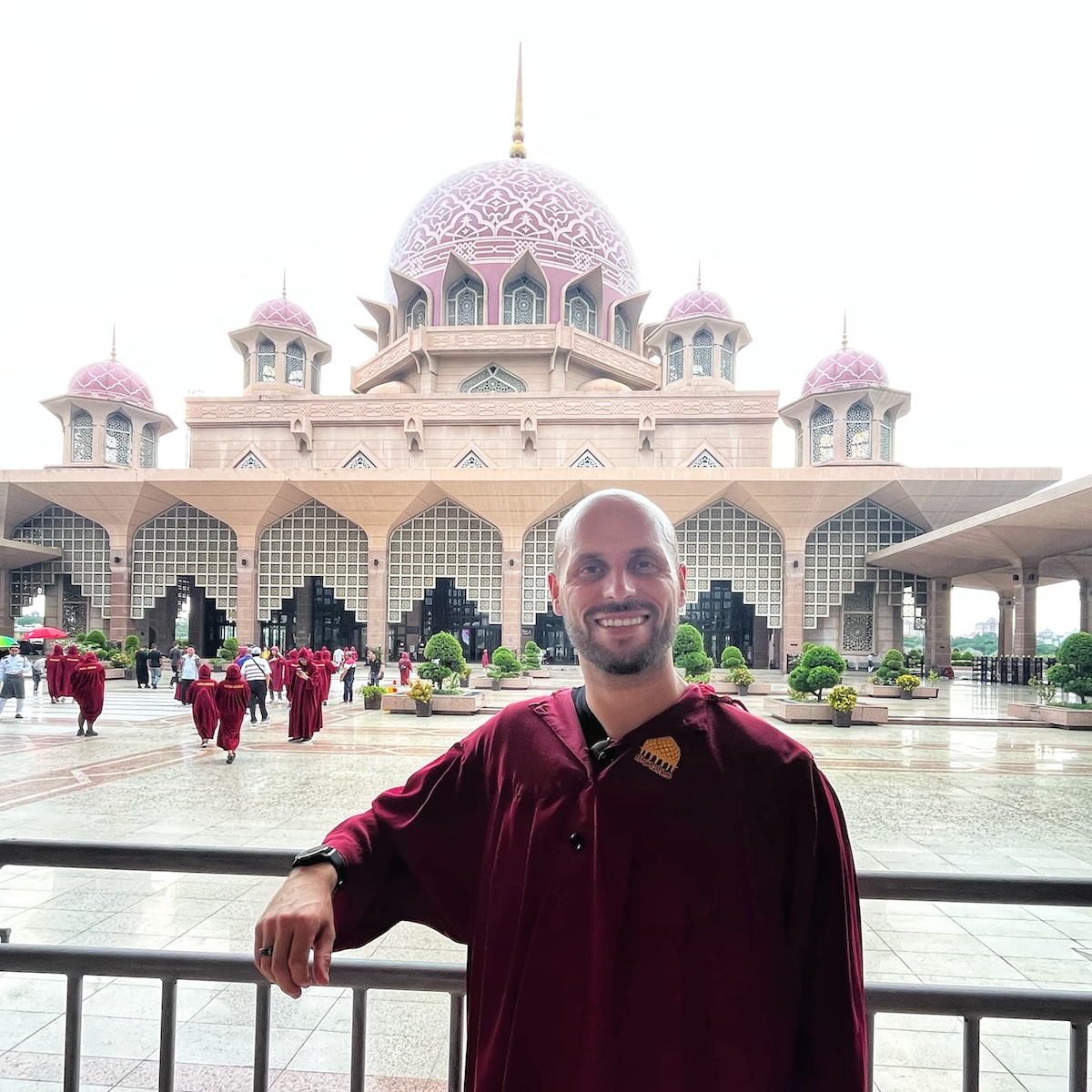 A man wearing a maroon robe standing in front of the Putra Mosque in Putrajaya, Malaysia, with its distinctive pink domes and intricate architectural design. Other visitors in similar robes can be seen in the background, highlighting the mosque's grandeur and cultural significance.