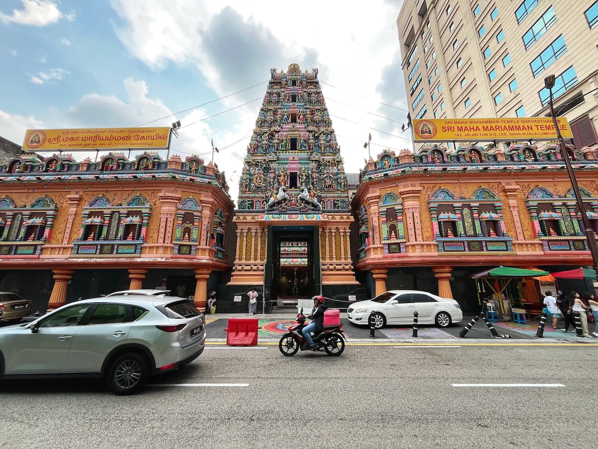Colourful and ornate Sri Maha Mariamman Temple in Kuala Lumpur, Malaysia, with detailed sculptures and vibrant decorations, flanked by city buildings. Motorists pass by on the street in front of the temple